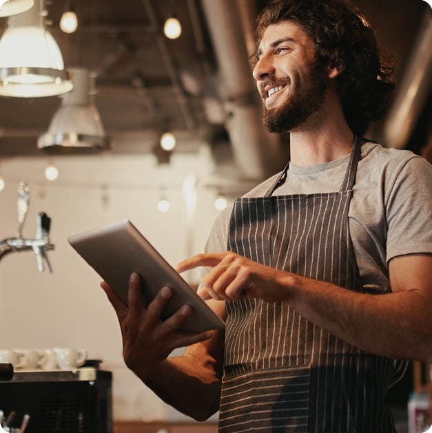 Smiling caucasian man wearing apron standing behind cafe counter using digital tablet looking away; Shutterstock ID 1728356797; purchase_order: -; job: -; client: -; other: -